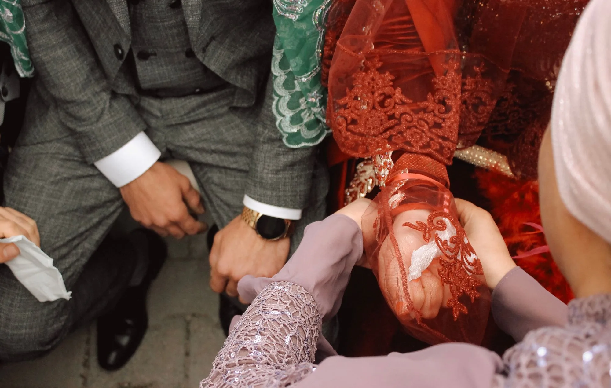 Kına gecesinde kırmızı duvaklı gelinin elini tutan yakınları ve diz çöken damat
