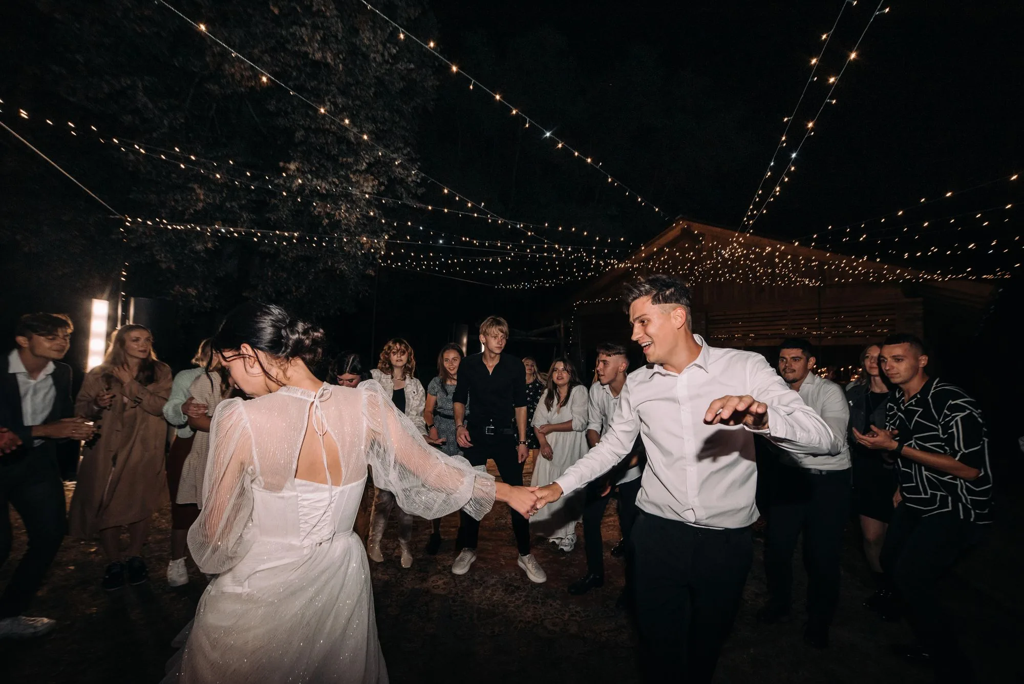 Gece ışık zincirleri altında misafirlerin arasında dans eden gelin ve damat